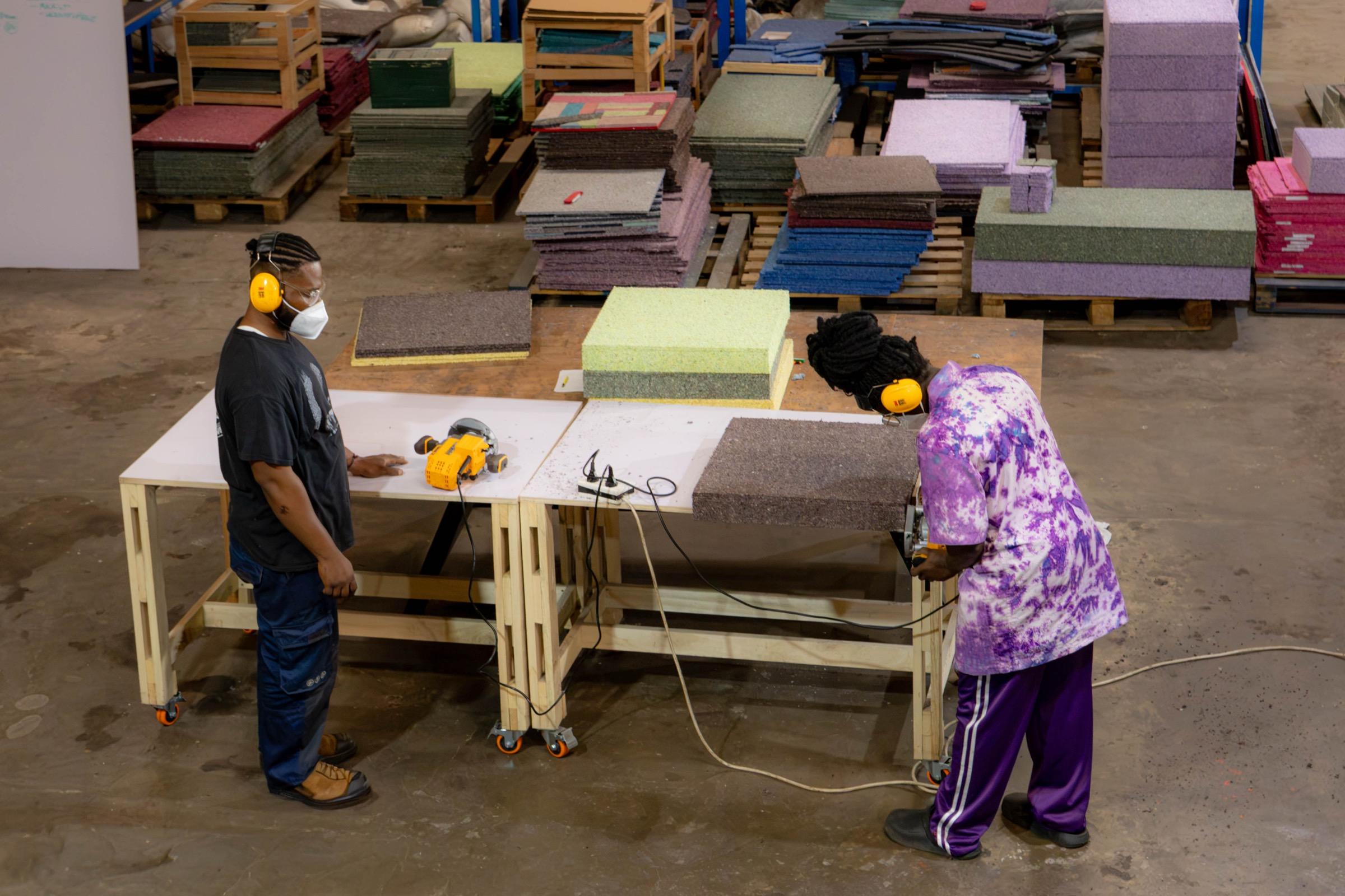 Two workers in ear defenders cutting recycled fibreboard panels at a workbench, with stacks of coloured board behind them.
